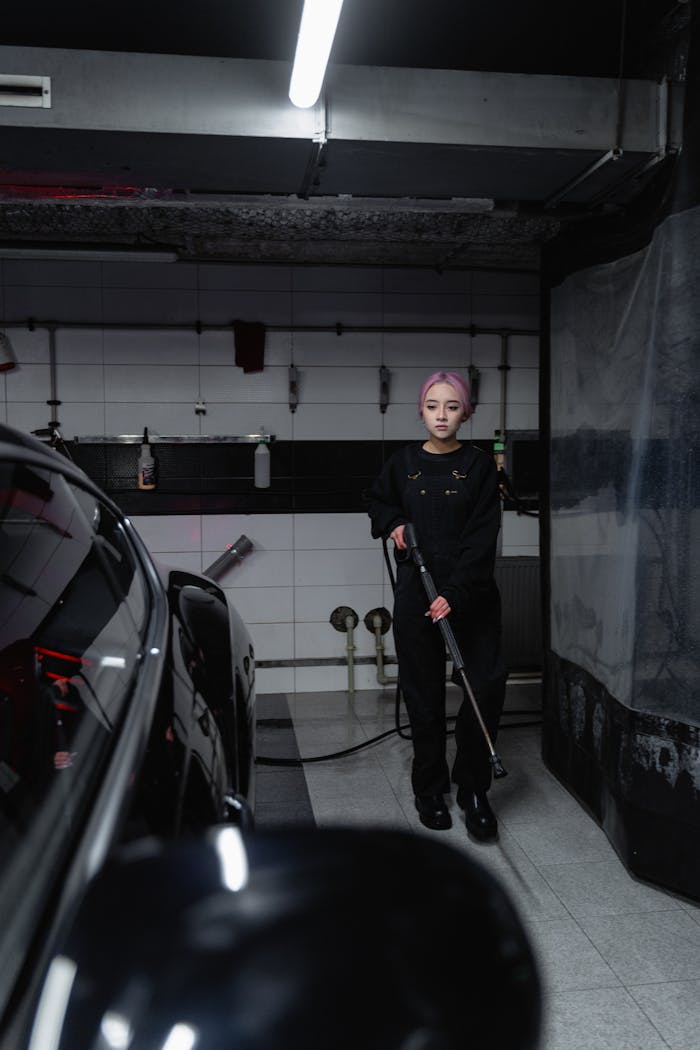 Asian woman uses pressure washer to clean car in garage with industrial vibe.