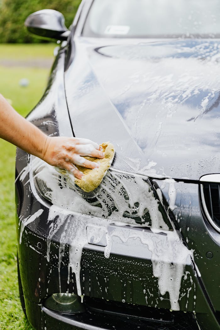 Person washing a black cars headlight with a sponge and soap outdoors.